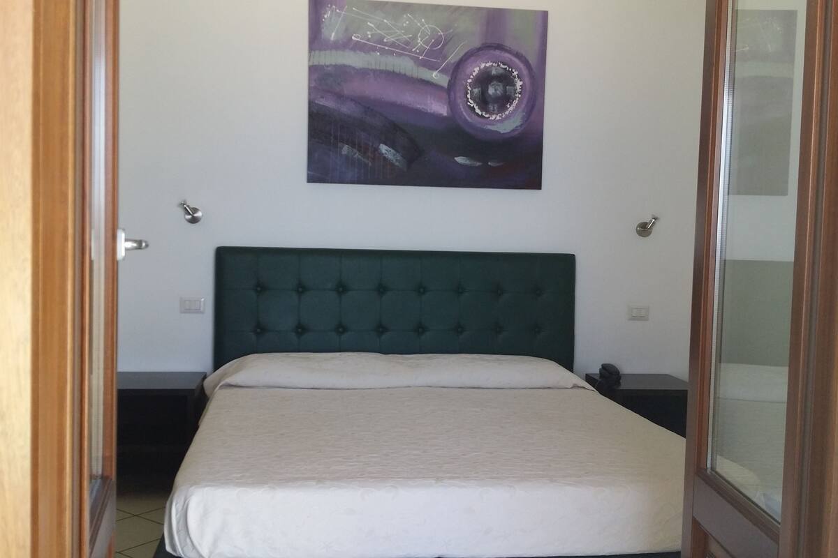 A bedroom with a green tufted headboard, a white bedspread, and a wall-mounted abstract painting.