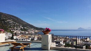 A terrace with a wooden lounge chair, a flower pot, and a view of a coastal town with buildings and a mountain in the background. - La Zagara Hotel (Lipari)