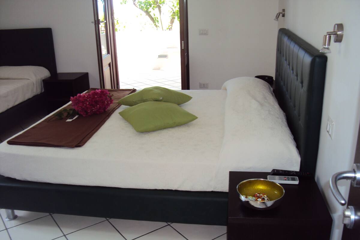 A neatly made bed with green pillows, a brown towel, and a pink flower on a white bedspread. There is a small table beside the bed with a bowl and a remote control.