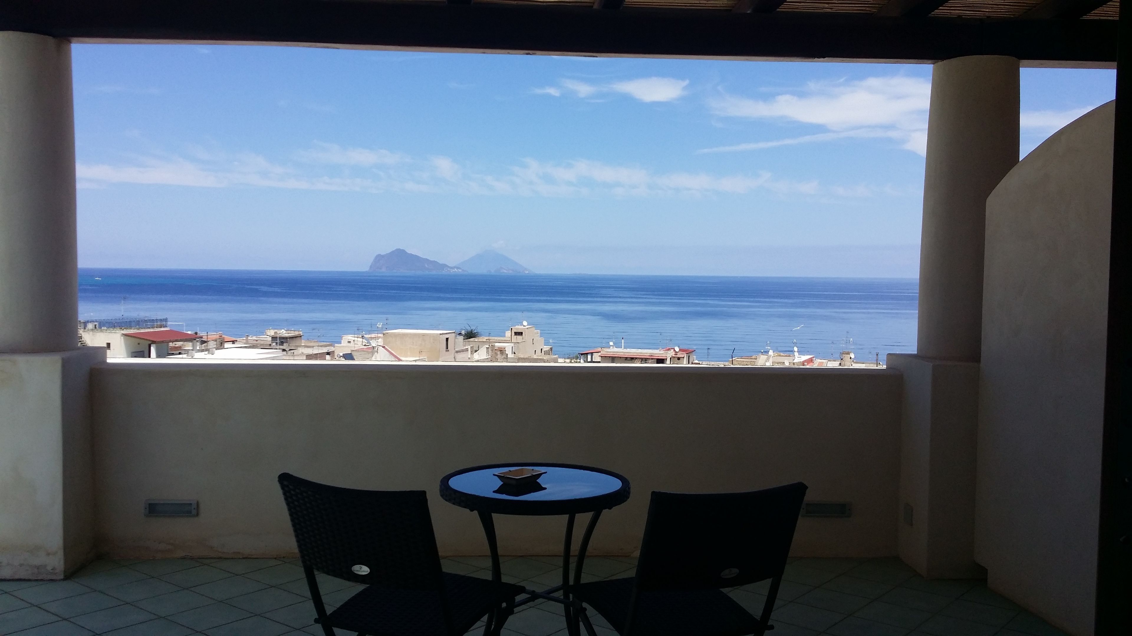 A balcony with two chairs and a small table, overlooking a coastal view with buildings and a distant mountain.