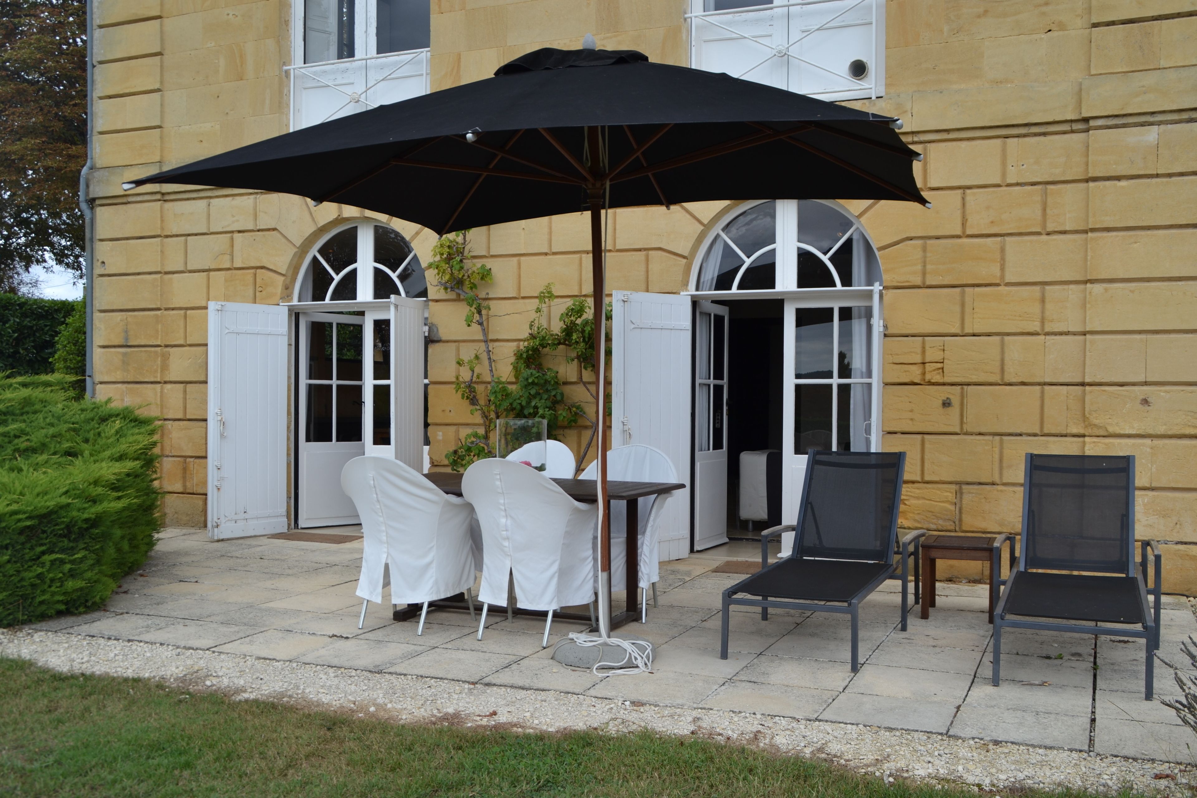 An outdoor patio with a table and chairs, a large black umbrella, and a building with arched windows and a door.