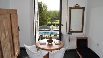 A room with a round wooden table, white chairs, a view of a pool and greenery outside, a mirror on the wall, and a radiator.