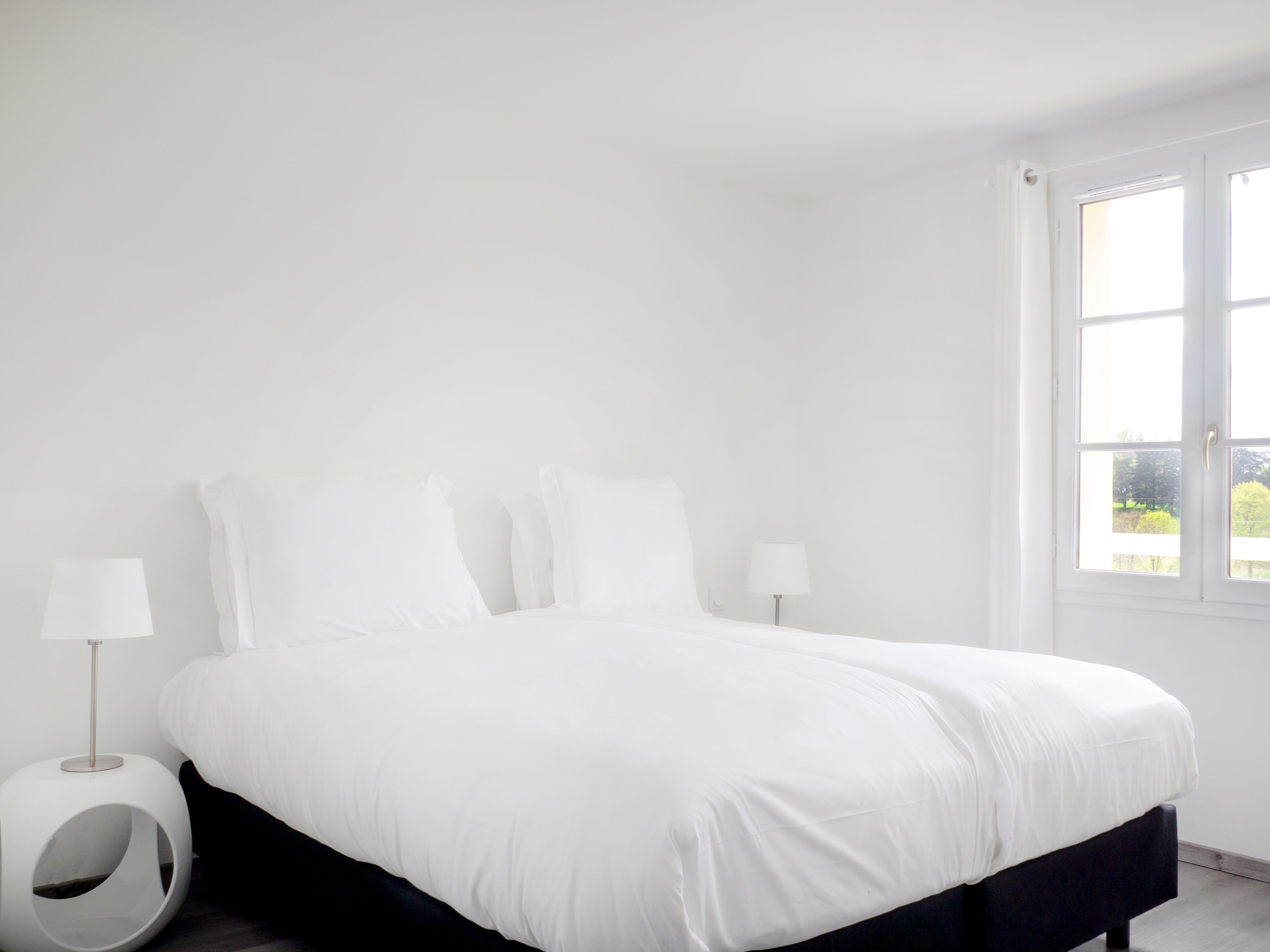 A minimalist bedroom with a large bed, two bedside lamps, and a window with a view of greenery.