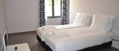 A neatly made bed with white linens and pillows, a bedside table with a remote control, and a window with curtains.