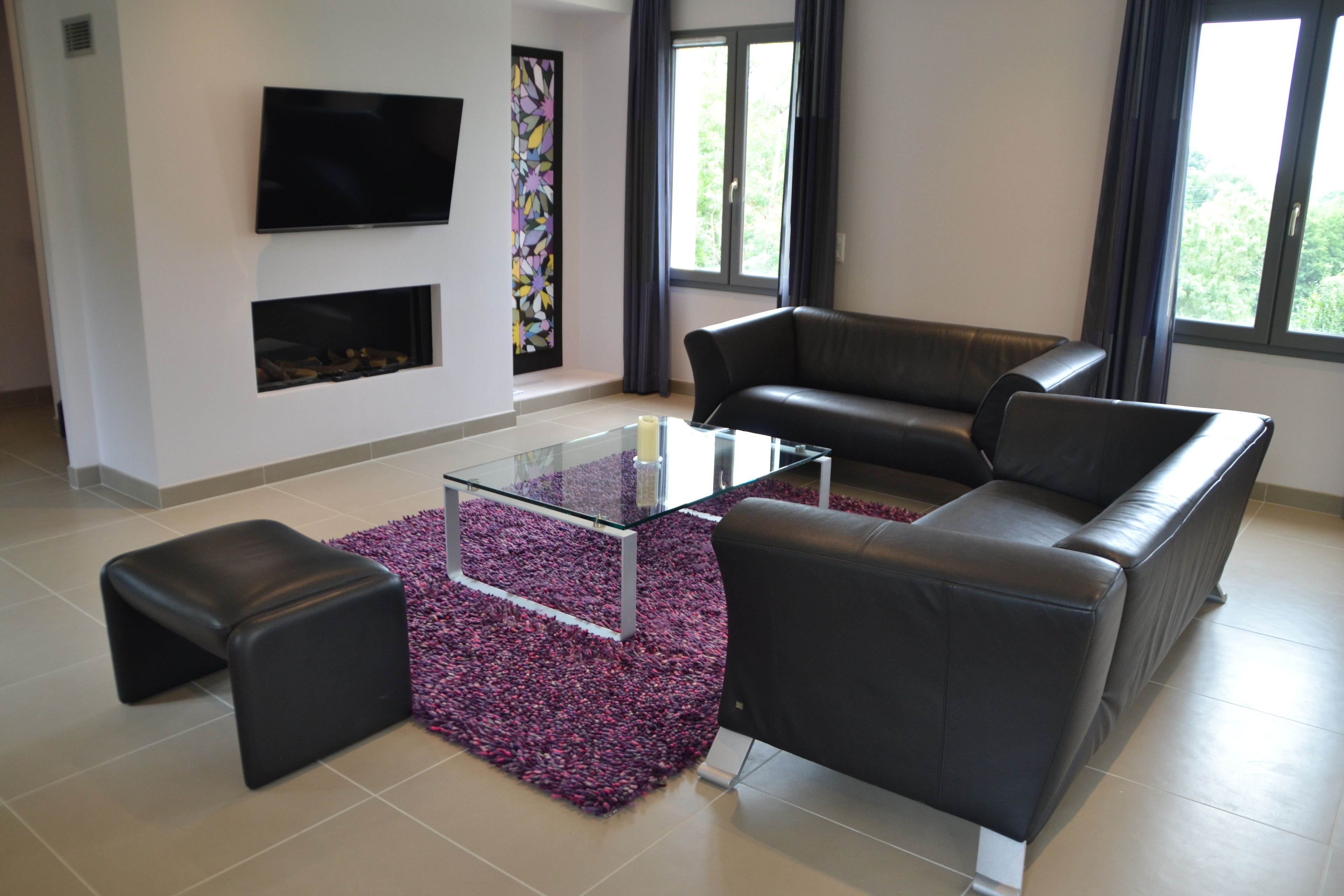 A modern living room with a glass coffee table, black leather sofa, and a flat-screen TV mounted on the wall.