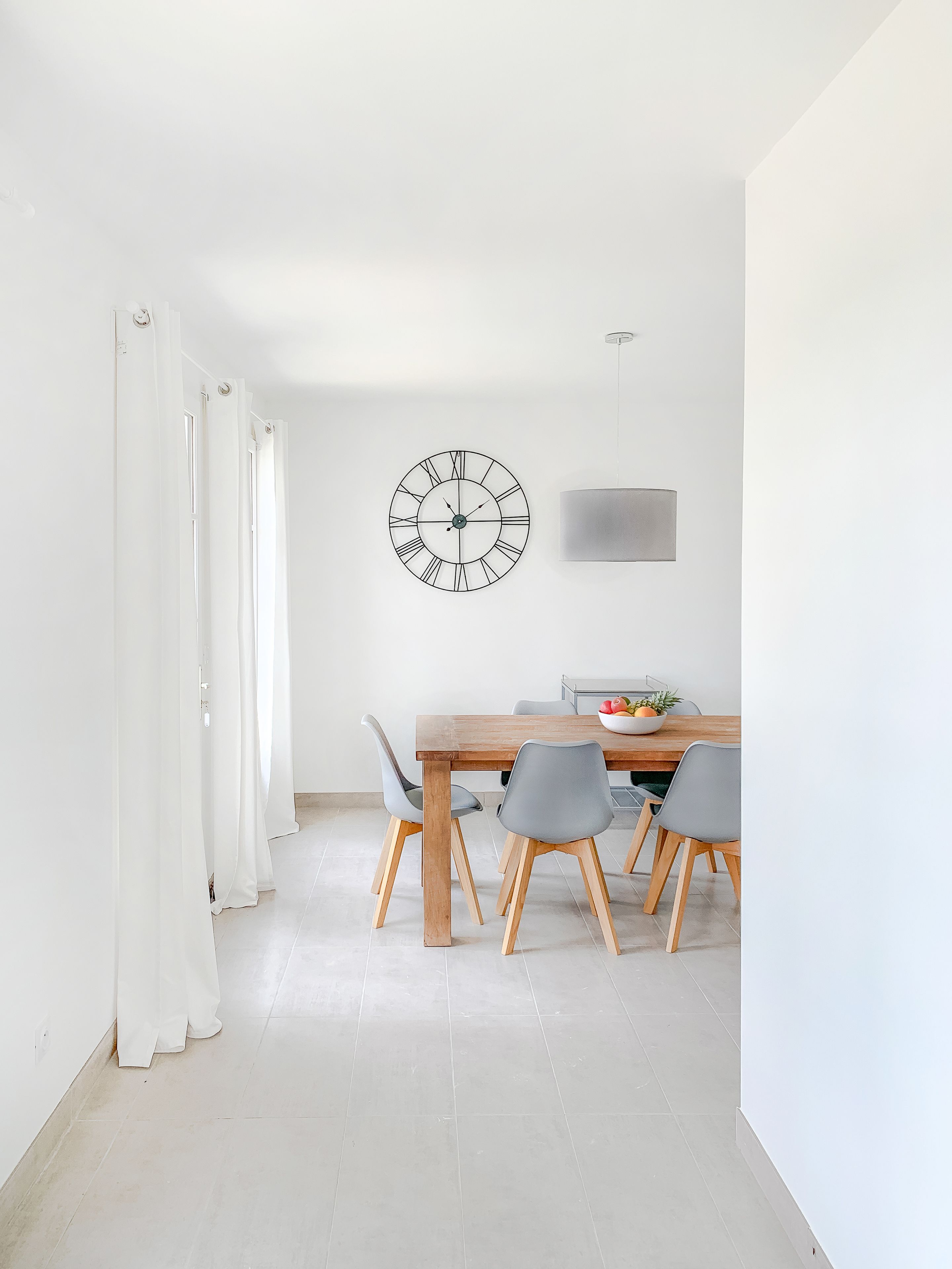 Un comedor con una mesa de madera, sillas grises, un reloj en la pared y una cortina blanca.