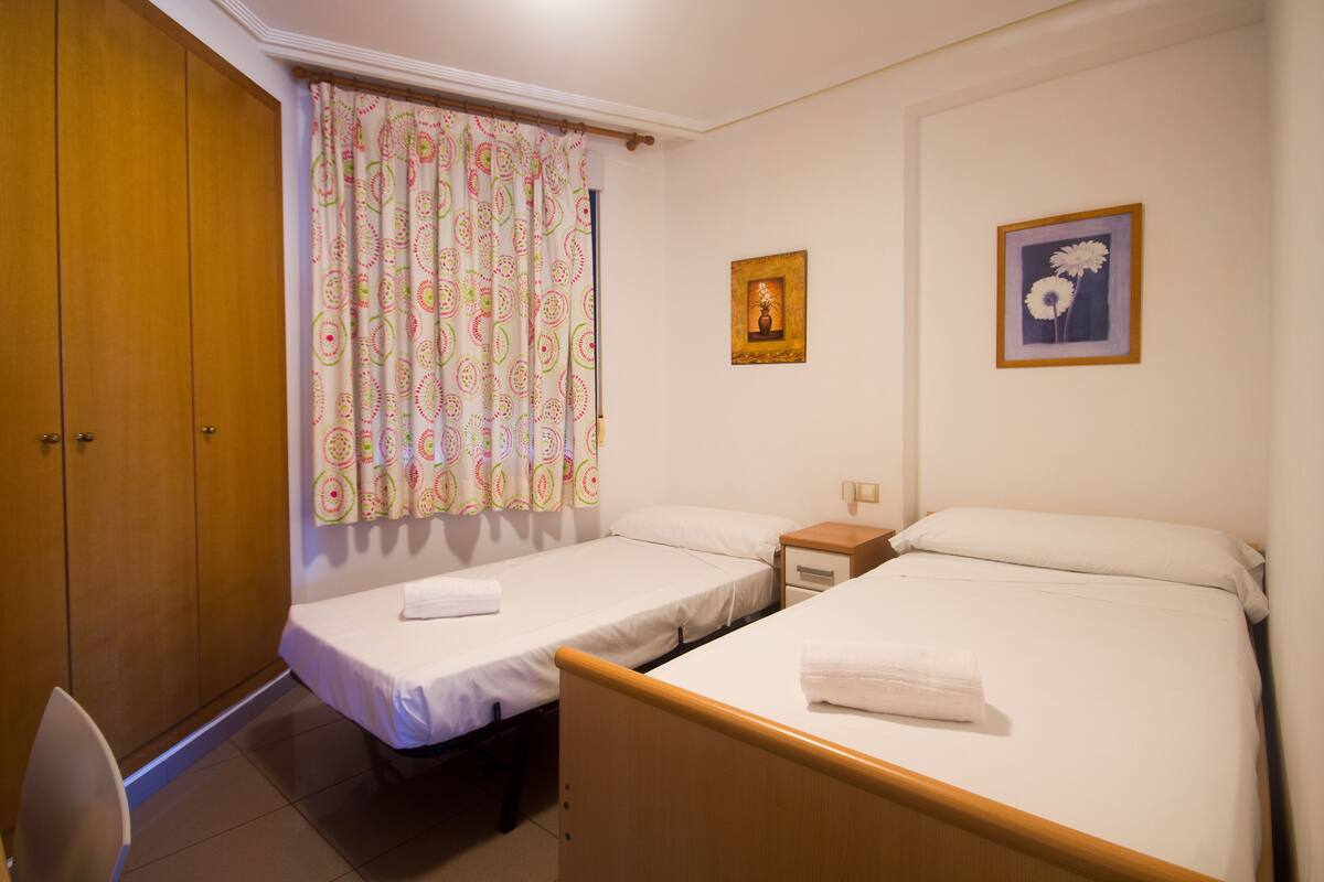 A room with two single beds, a wooden wardrobe, a small bedside table, and a curtain with a colorful pattern.