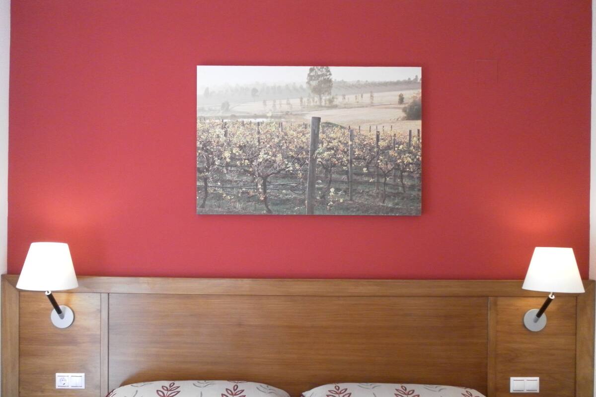 A hotel room with a red wall, a framed picture of a vineyard, two bedside lamps, and a bed with patterned pillows.