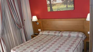 A double bed with a leaf-patterned bedspread, a wooden headboard, a nightstand with a lamp, a painting of trees on the wall, and a window with curtains.