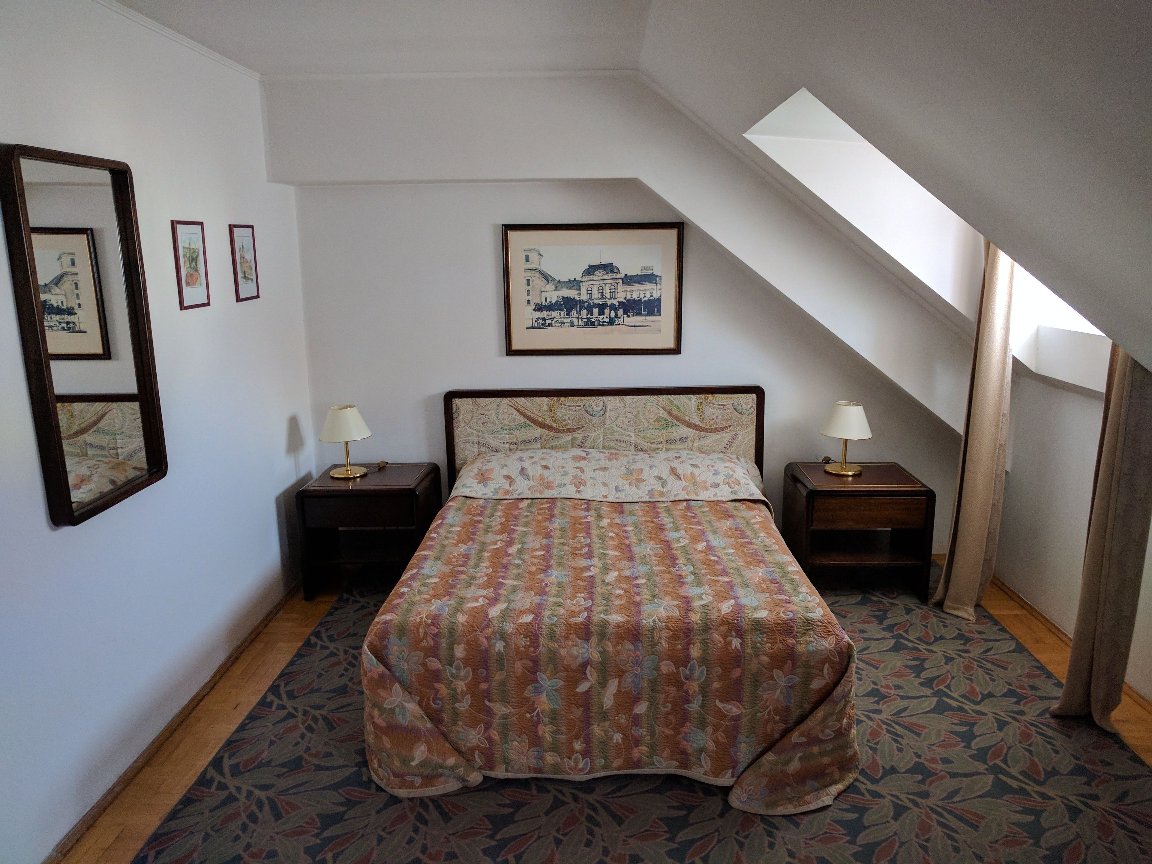 A bedroom with a bed, two bedside tables, a mirror, framed pictures, and a skylight.