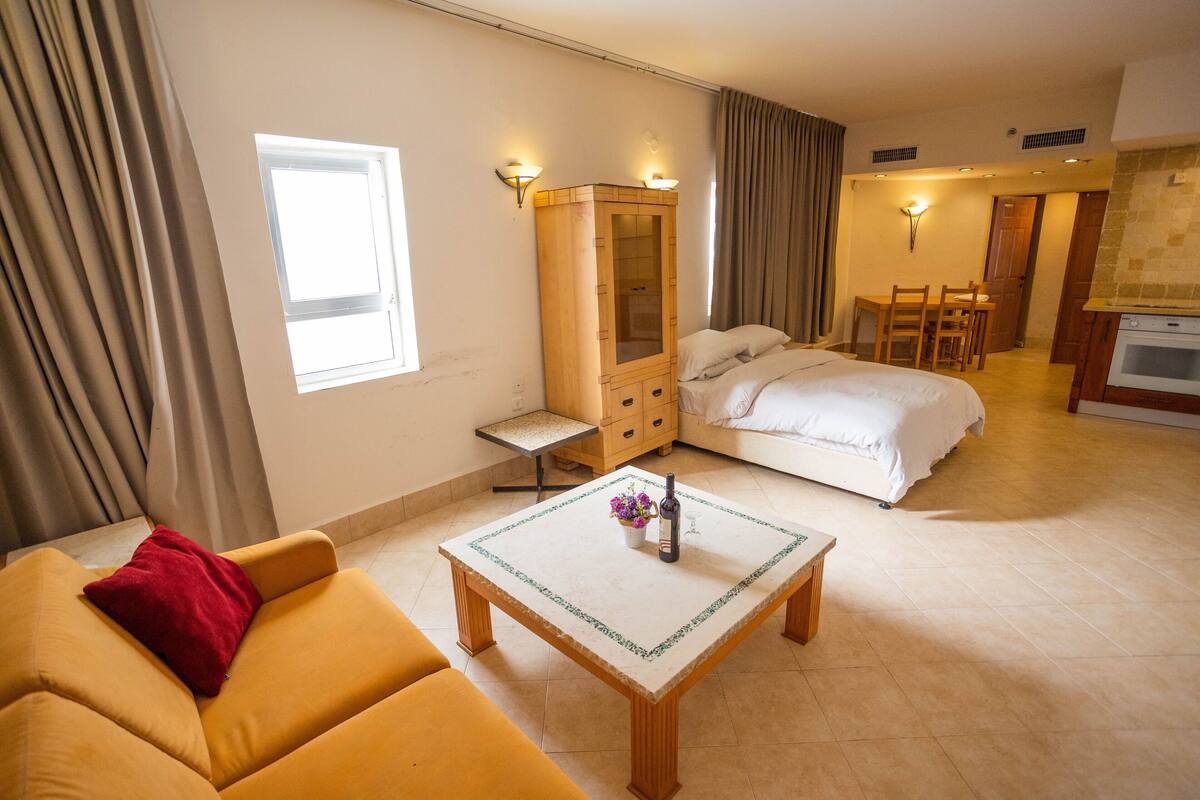 A hotel room with a bed, a sofa, a small table with a bottle and flowers, a wardrobe, and a kitchenette.