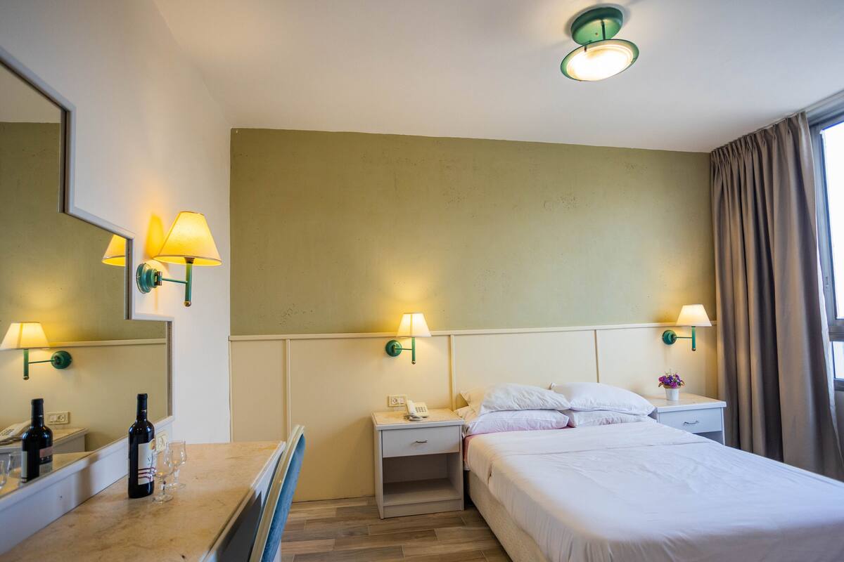 A hotel room with a bed, bedside tables, a mirror, and a window with curtains.