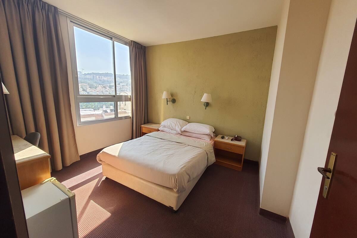 A hotel room with a large bed, a desk, a chair, a nightstand with a lamp, and a view of the city through the window.