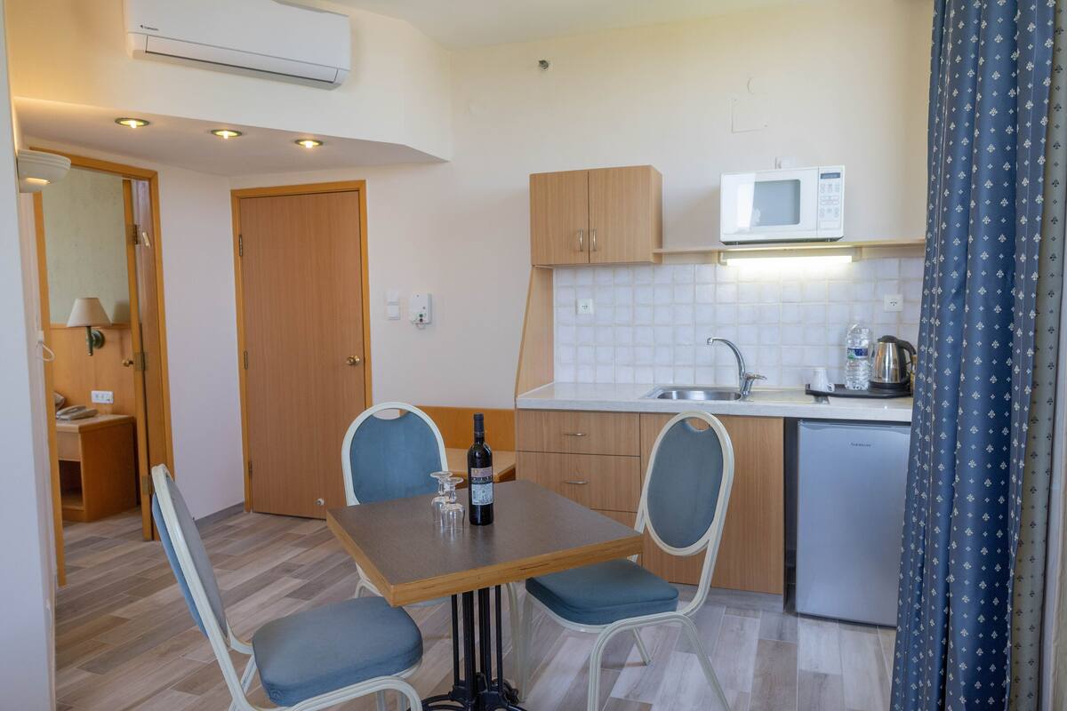 A compact kitchen with a microwave, sink, and a small refrigerator. There is a dining table with chairs and a bottle of wine on it.