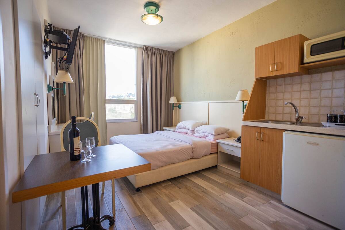 A compact hotel room with a bed, a small kitchenette, a dining table, and a window with curtains.