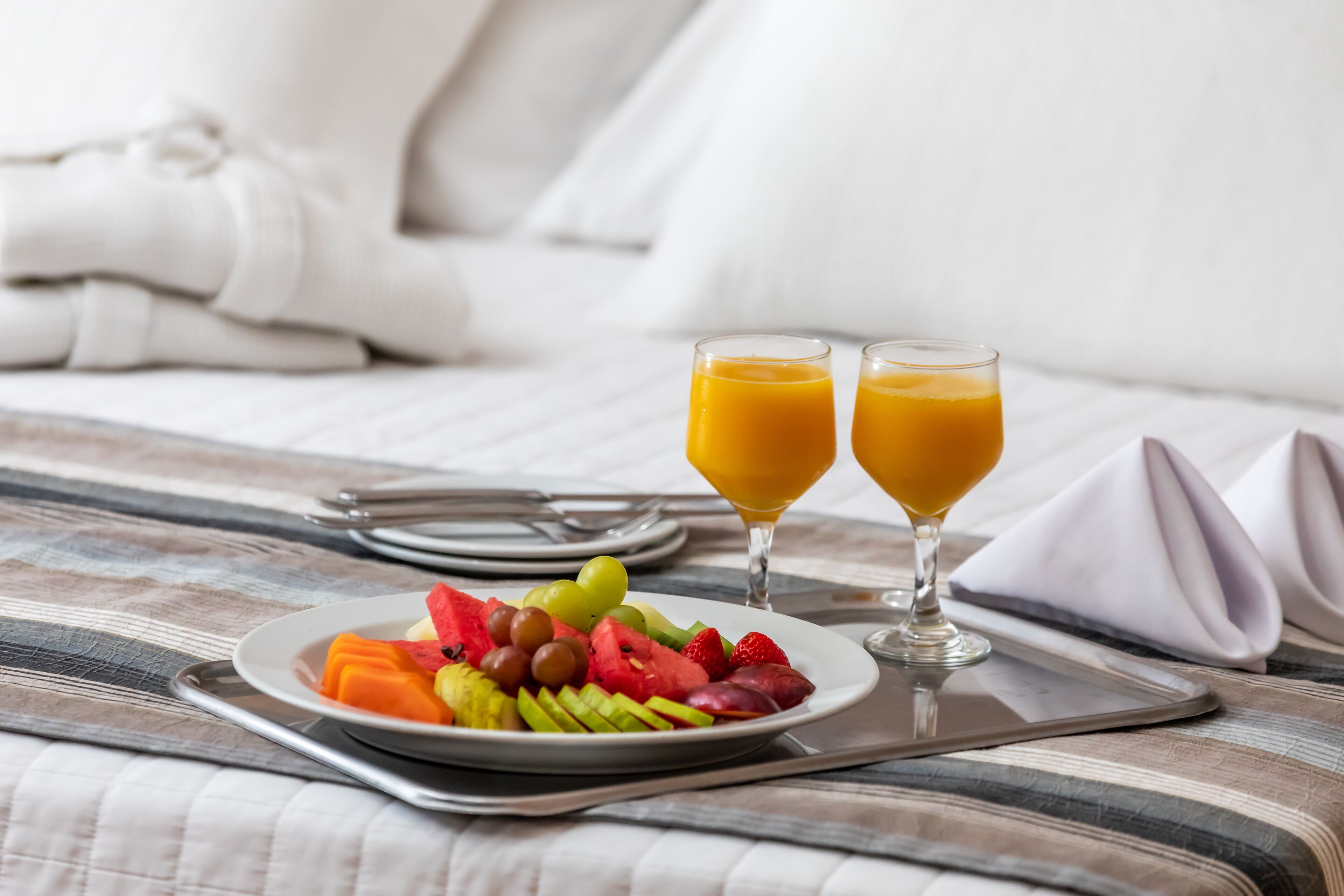 Desayuno con un plato de fruta y dos vasos de zumo de naranja sobre una cama.