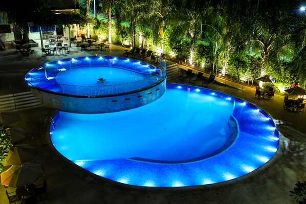 A circular pool with a smaller inner pool, surrounded by lounge chairs and tables, with trees and ambient lighting in the background.