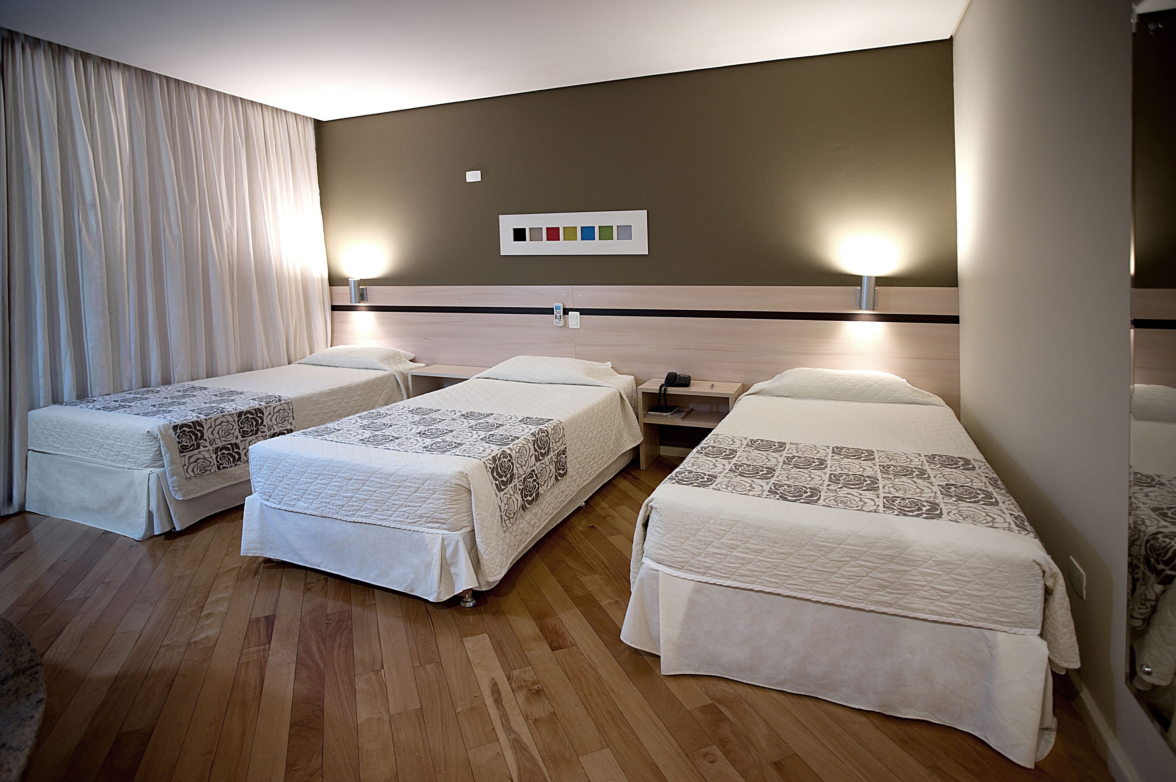 A hotel room with three single beds, wooden flooring, a large window with curtains, and a wall-mounted light switch.