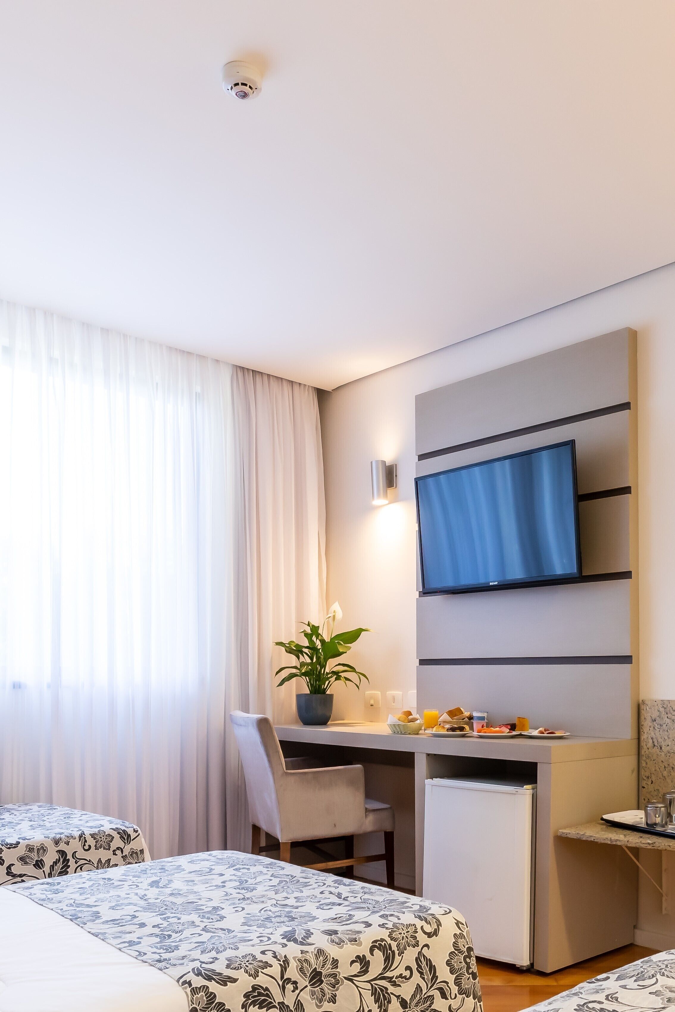 A hotel room with two beds, a desk with a TV, a small fridge, and a potted plant.