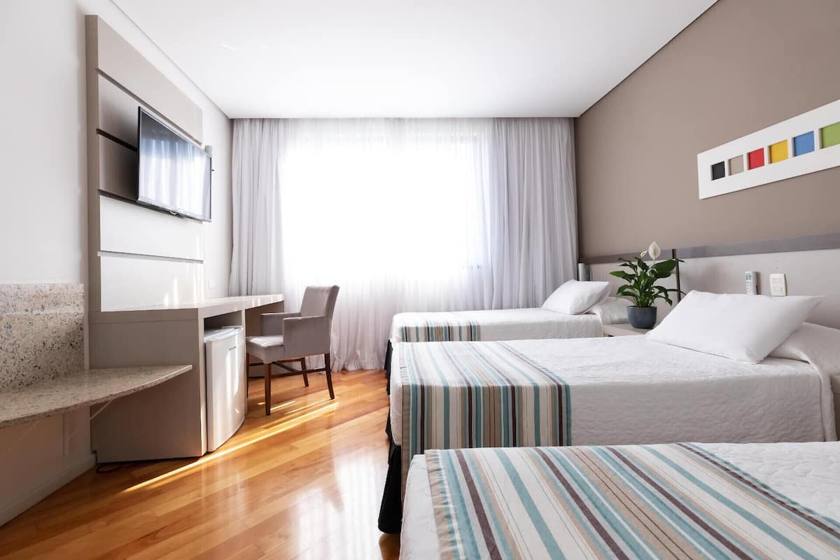 Quarto de hotel com cama, escrivaninha, cadeira e televisão.