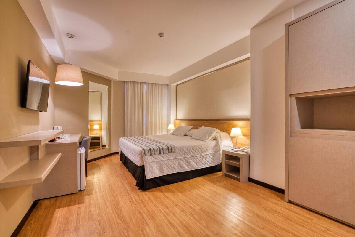 A hotel room with a bed, bedside tables, a TV, a mirror, a wardrobe, and a window with curtains.