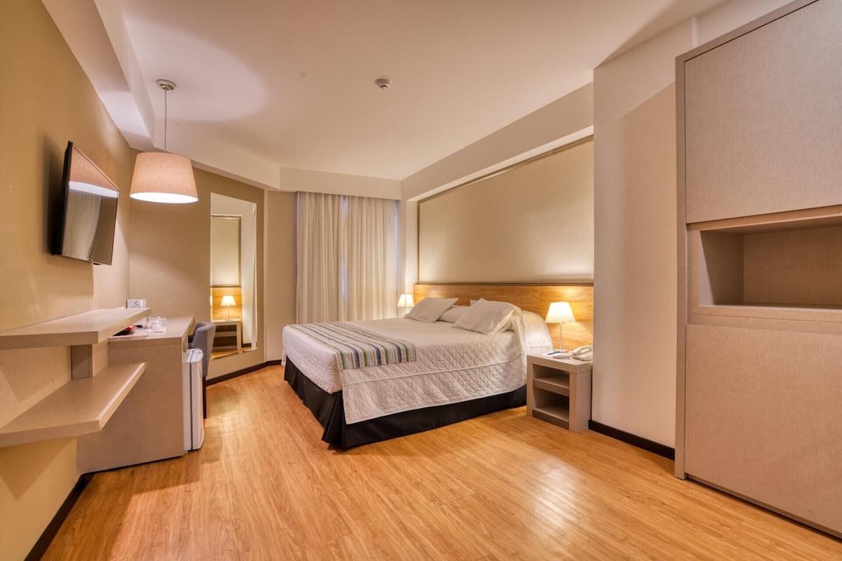Quarto de hotel com cama, mesas de cabeceira, televisão, espelho, guarda-roupa e janela com cortinas.