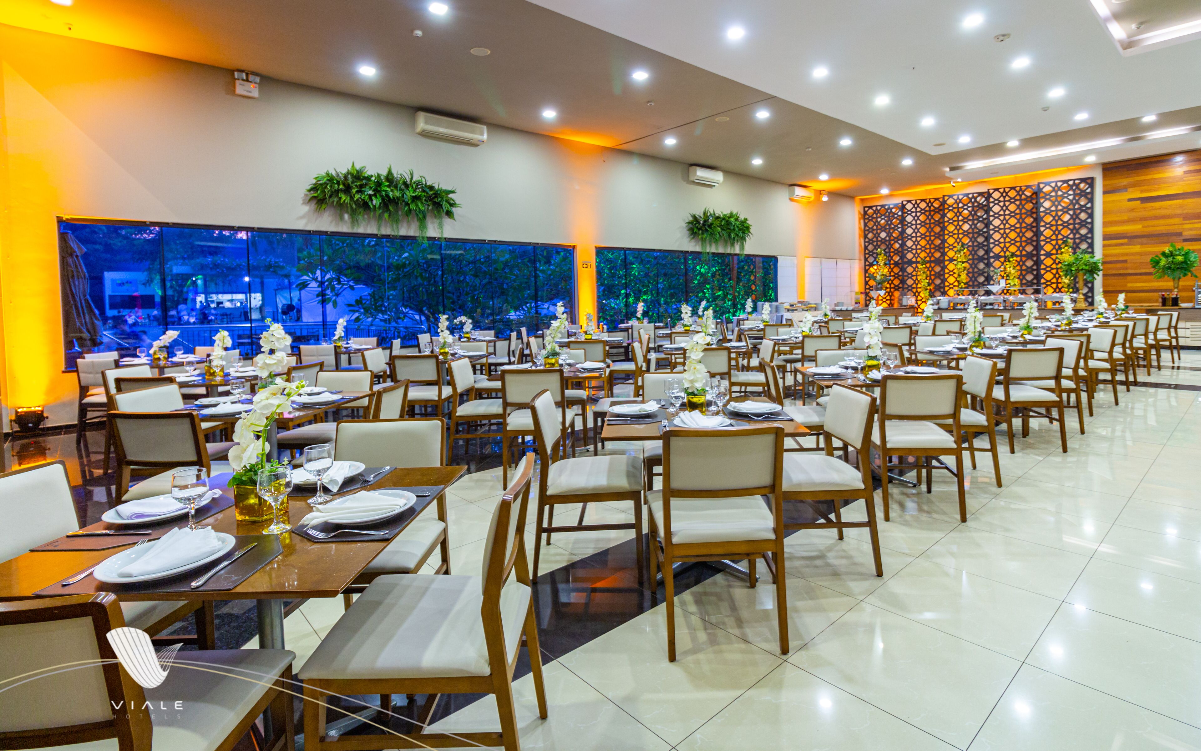 A spacious restaurant with multiple tables and chairs, air conditioning units, and decorative plants.