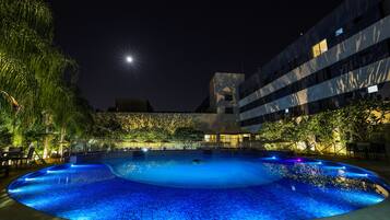 A nighttime swimming pool with a curved edge, surrounded by trees and a building with multiple windows.
