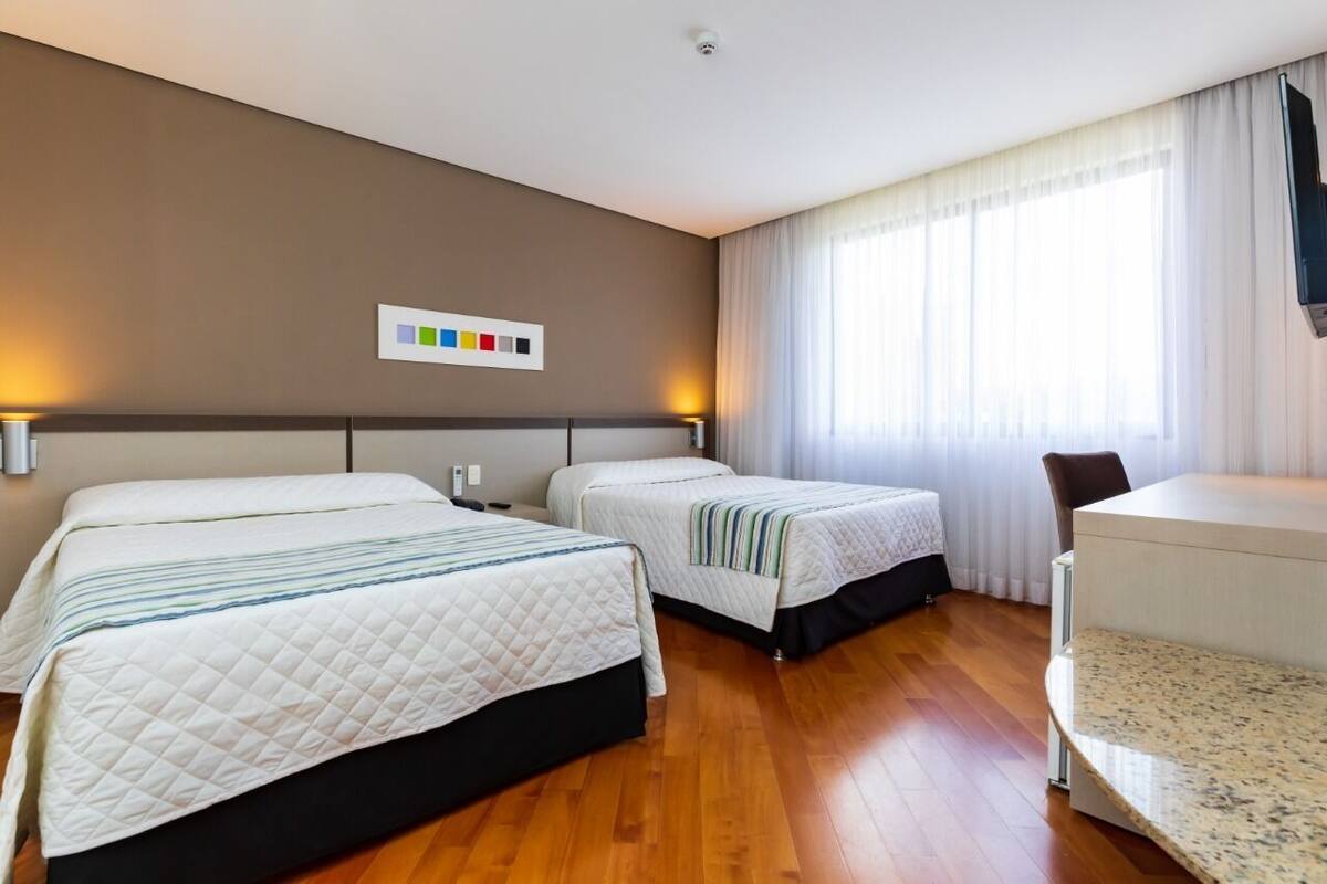Une chambre d’hôtel avec deux lits, un bureau et une chaise. Il y a une fenêtre avec des rideaux et un téléviseur fixé au mur.