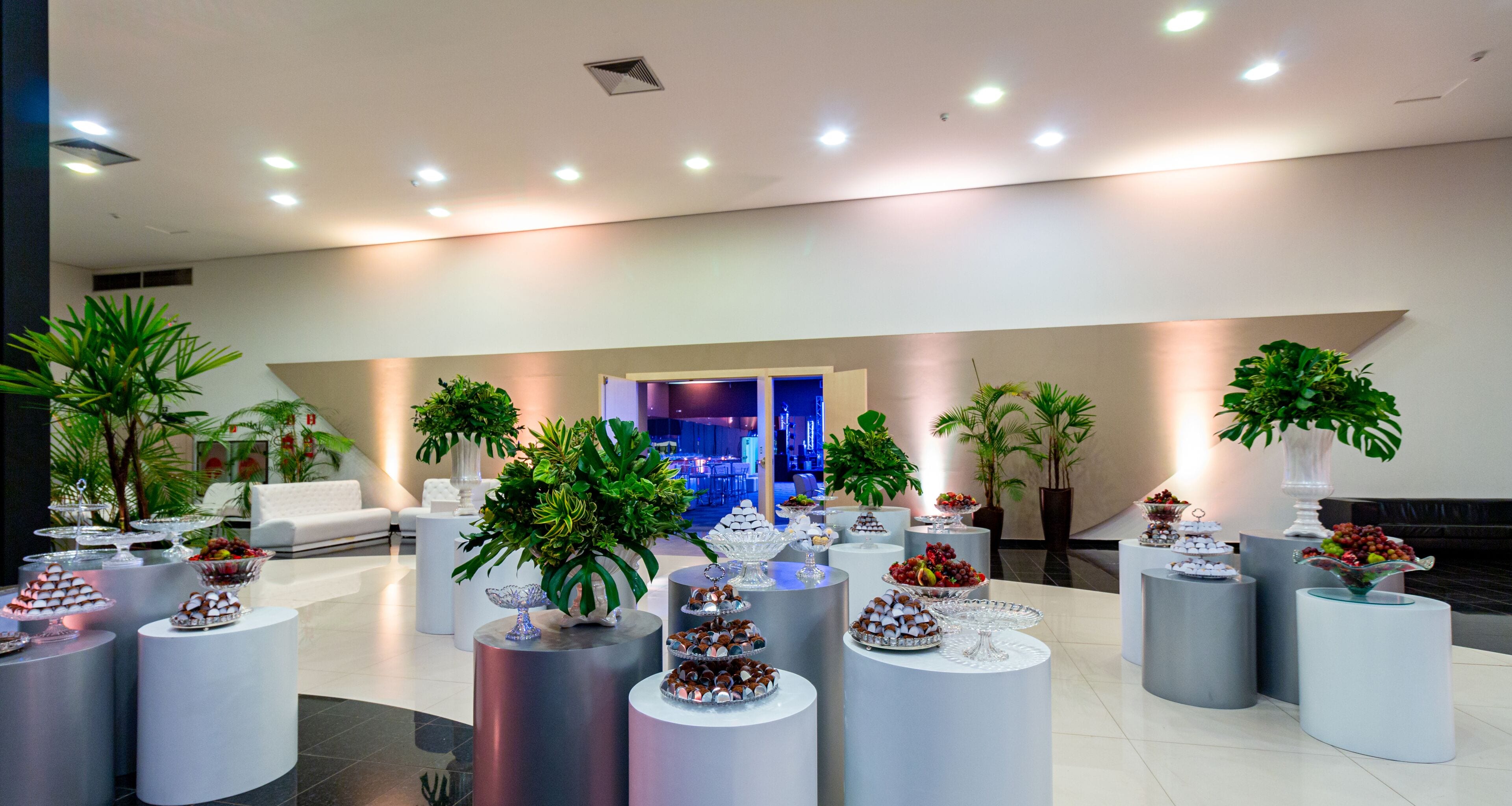 A modern lobby with round tables displaying various food items, surrounded by potted plants and a seating area with a white couch.