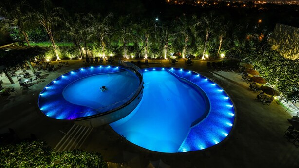 Vista nocturna de una piscina con diseño circular, rodeada de palmeras y sillas de descanso.