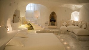 Quarto aconchegante com decoração em tons de branco, cama de linhas curvas, lareira e área de estar com almofadas.