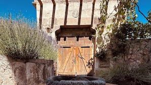 Uma fonte de pedra com água e flores num pátio com porta de madeira e paredes de pedra.
