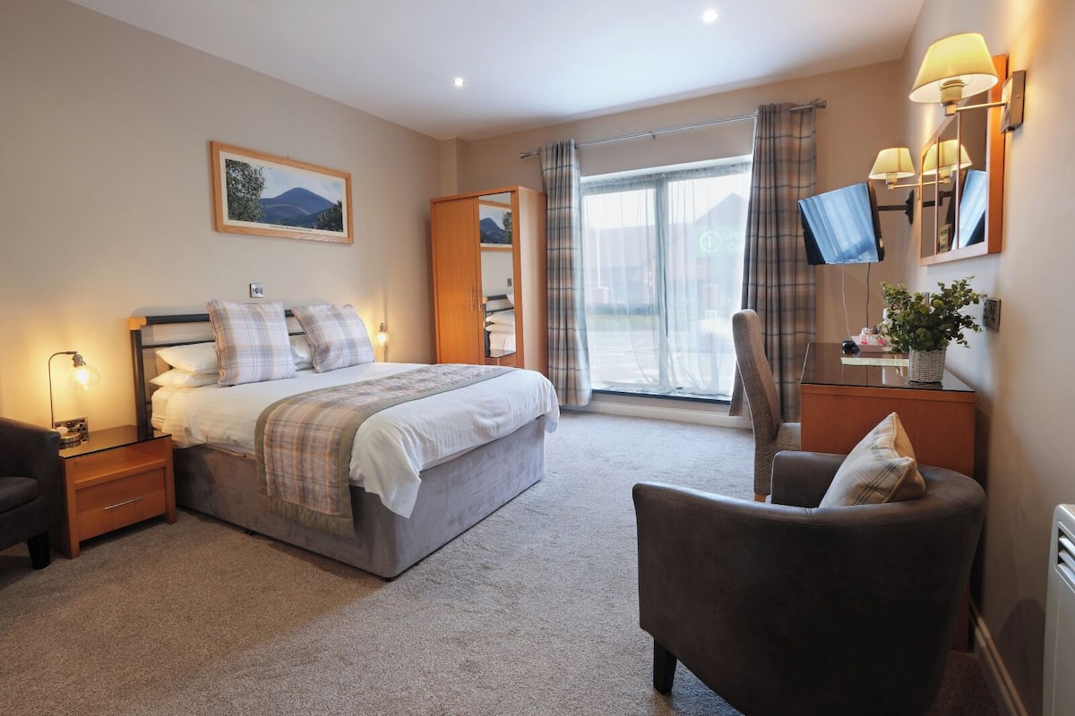 A hotel room with a bed, bedside table, lamp, chair, and a large window with curtains.