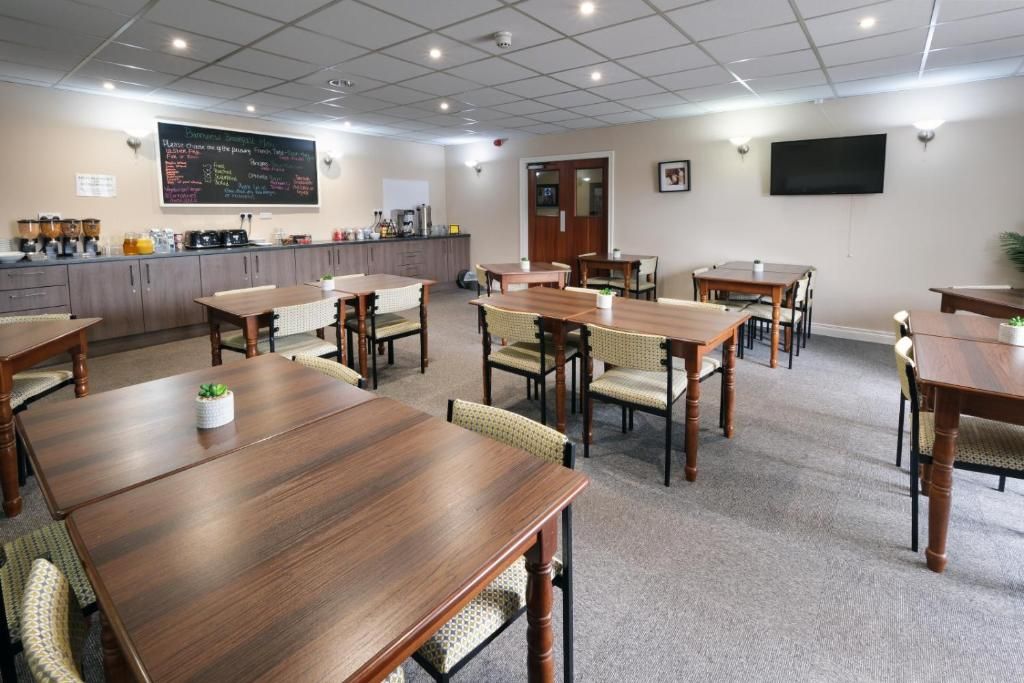 A conference room with multiple wooden tables and chairs, a kitchen area with coffee machines, and a chalkboard menu on the wall.