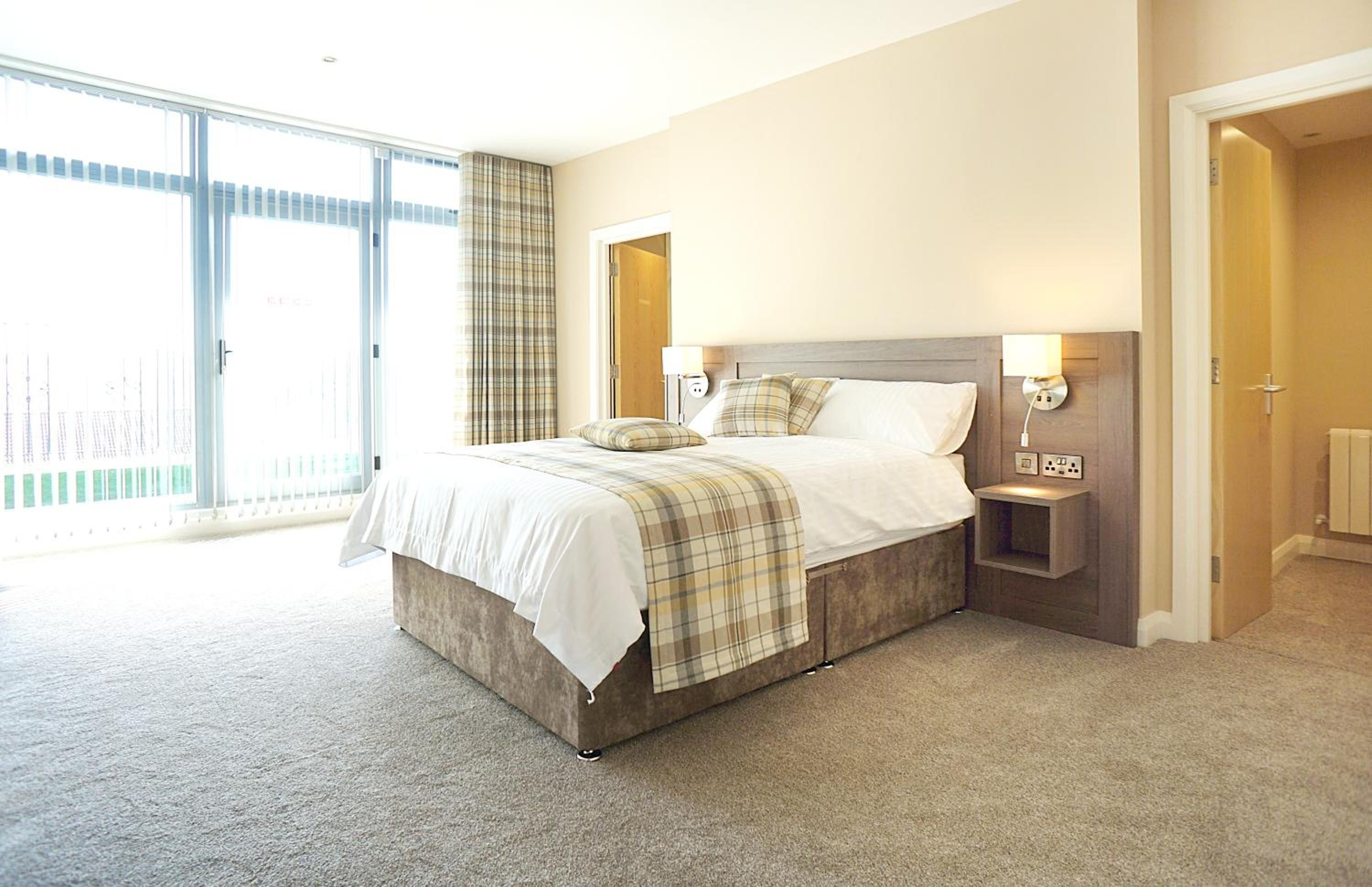 A bedroom with a large bed, a nightstand, a wall-mounted lamp, and a sliding glass door leading to an outdoor area.