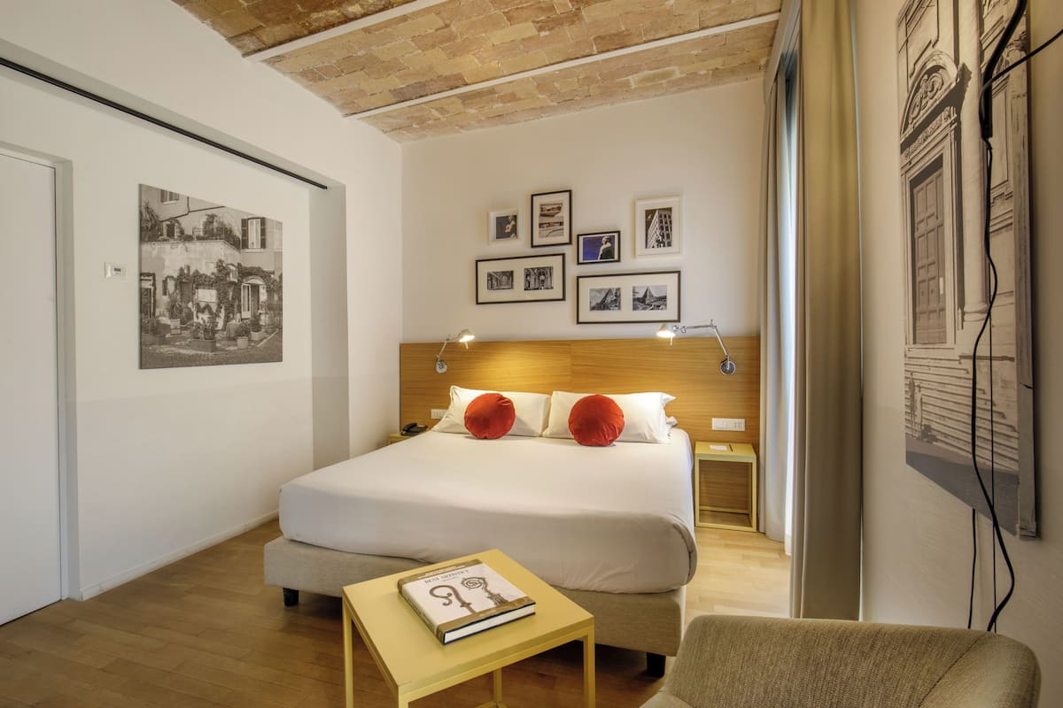 Una habitación de hotel con una cama grande, dos almohadas rojas, una mesita de noche con un libro y cuadros en la pared.