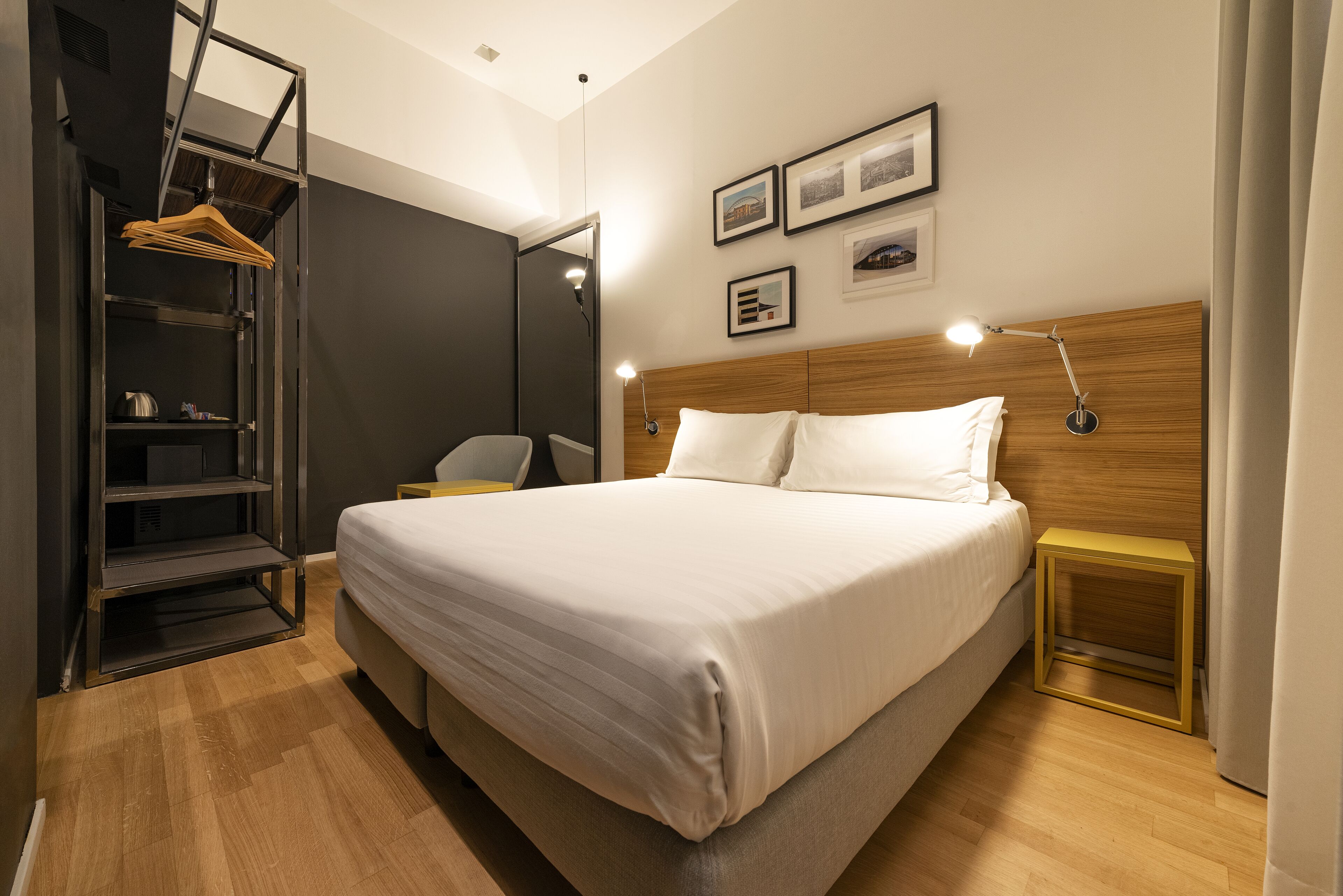 A modern hotel room with a large bed, a wooden headboard, bedside tables, a wardrobe with a mirror, and framed pictures on the wall.