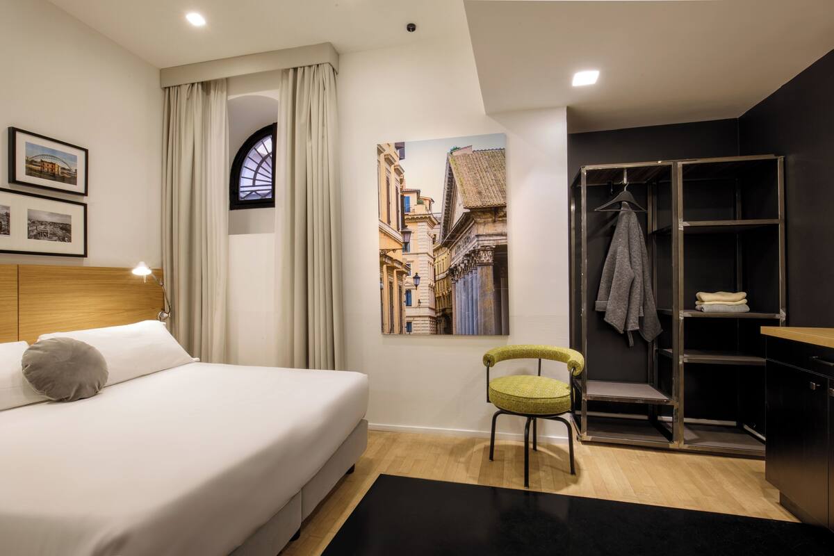 Una habitación de hotel moderna con cama, una silla, armario y un cuadro de una escena callejera.