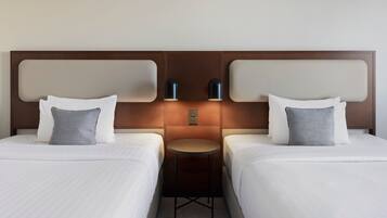 Habitación de hotel con dos camas, cada una con ropa de cama blanca y almohadas estampadas. Una pequeña mesa redonda entre las camas con dos lámparas fijadas a la pared.