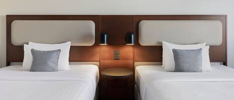 A hotel room with two beds, each with white bedding and patterned pillows. There is a small round table between the beds with two wall-mounted lamps.