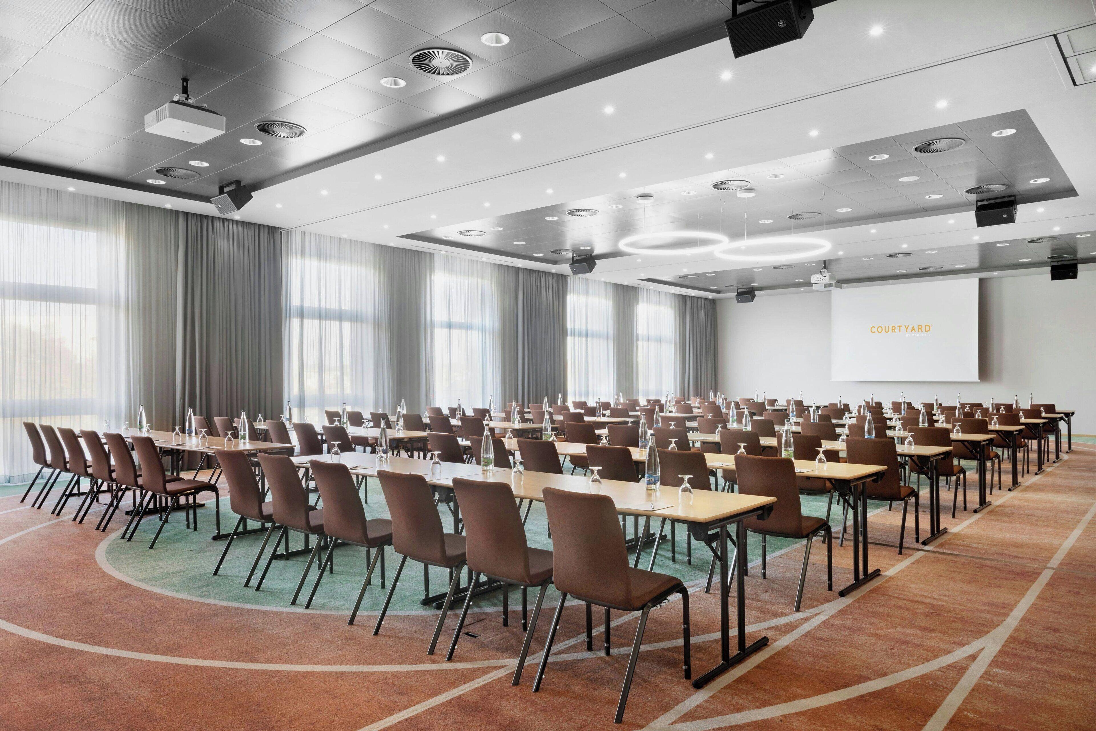 Une salle de conférence avec des rangées de tables et de chaises, un écran de projection et une grande fenêtre avec des rideaux.