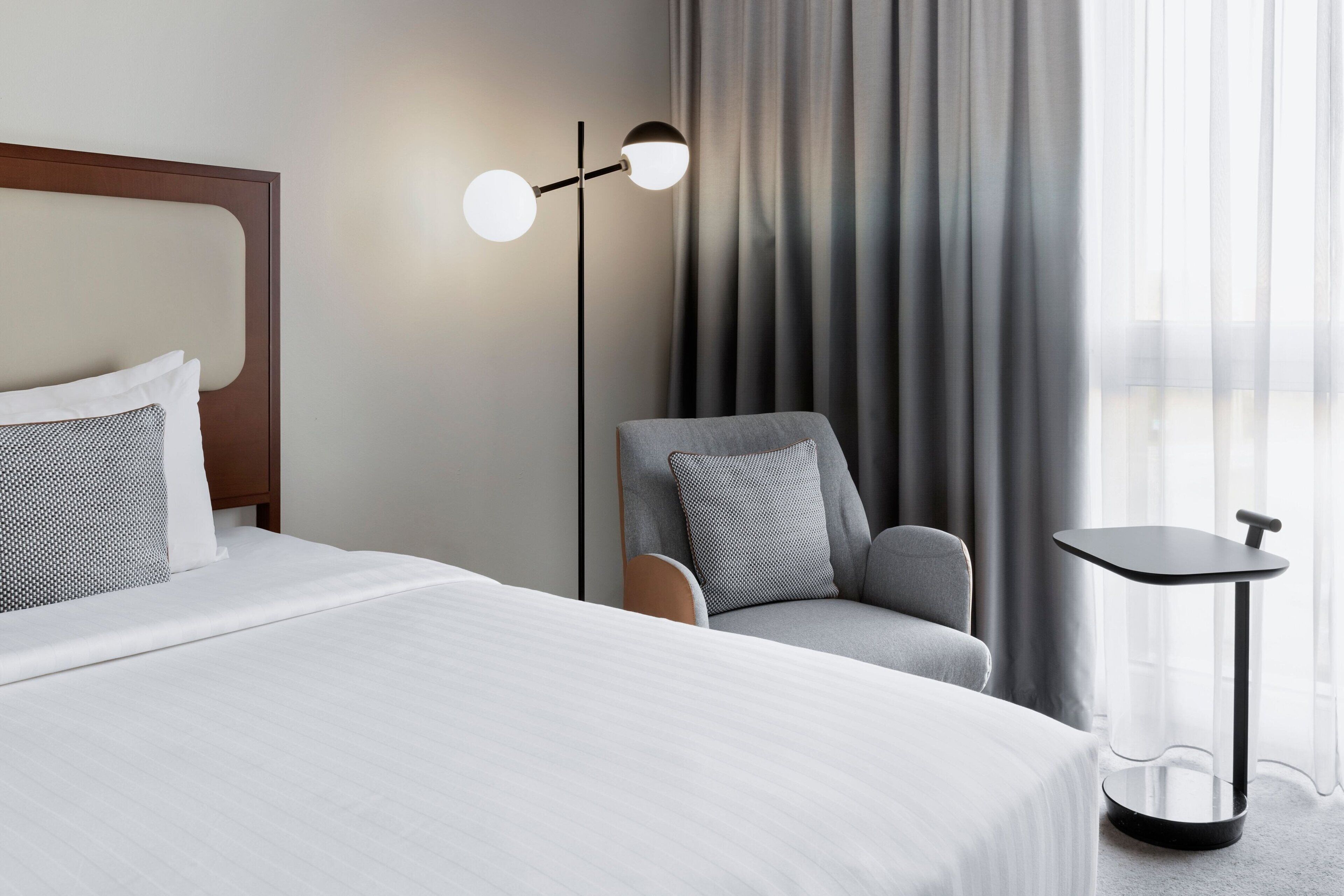 Ett hotellrum med en säng, en stol, ett litet bord och en golvlampa.