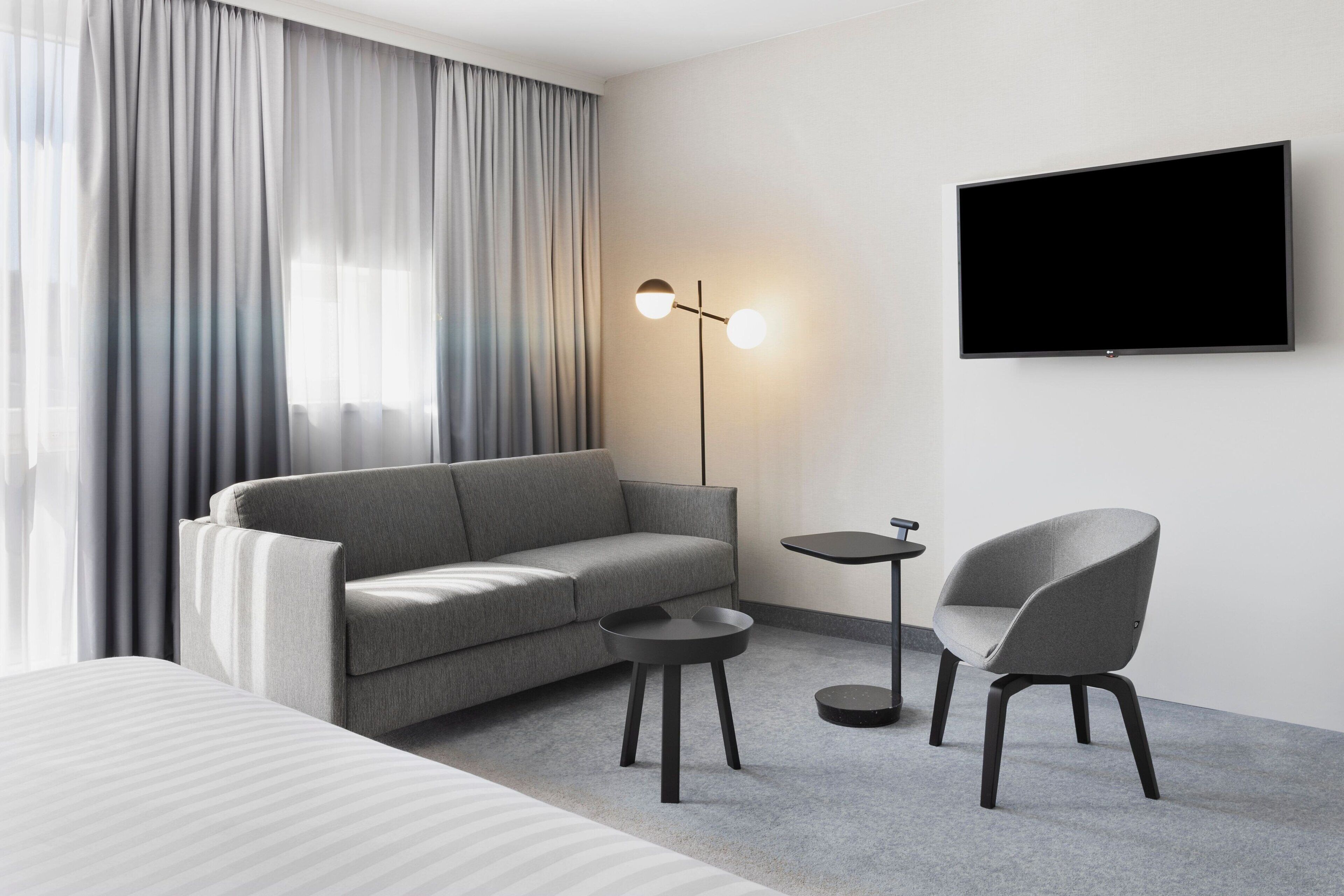 Une chambre d’hôtel moderne avec un canapé gris, une petite table ronde, un fauteuil gris, un téléviseur à écran plat et une lampe sur pied.