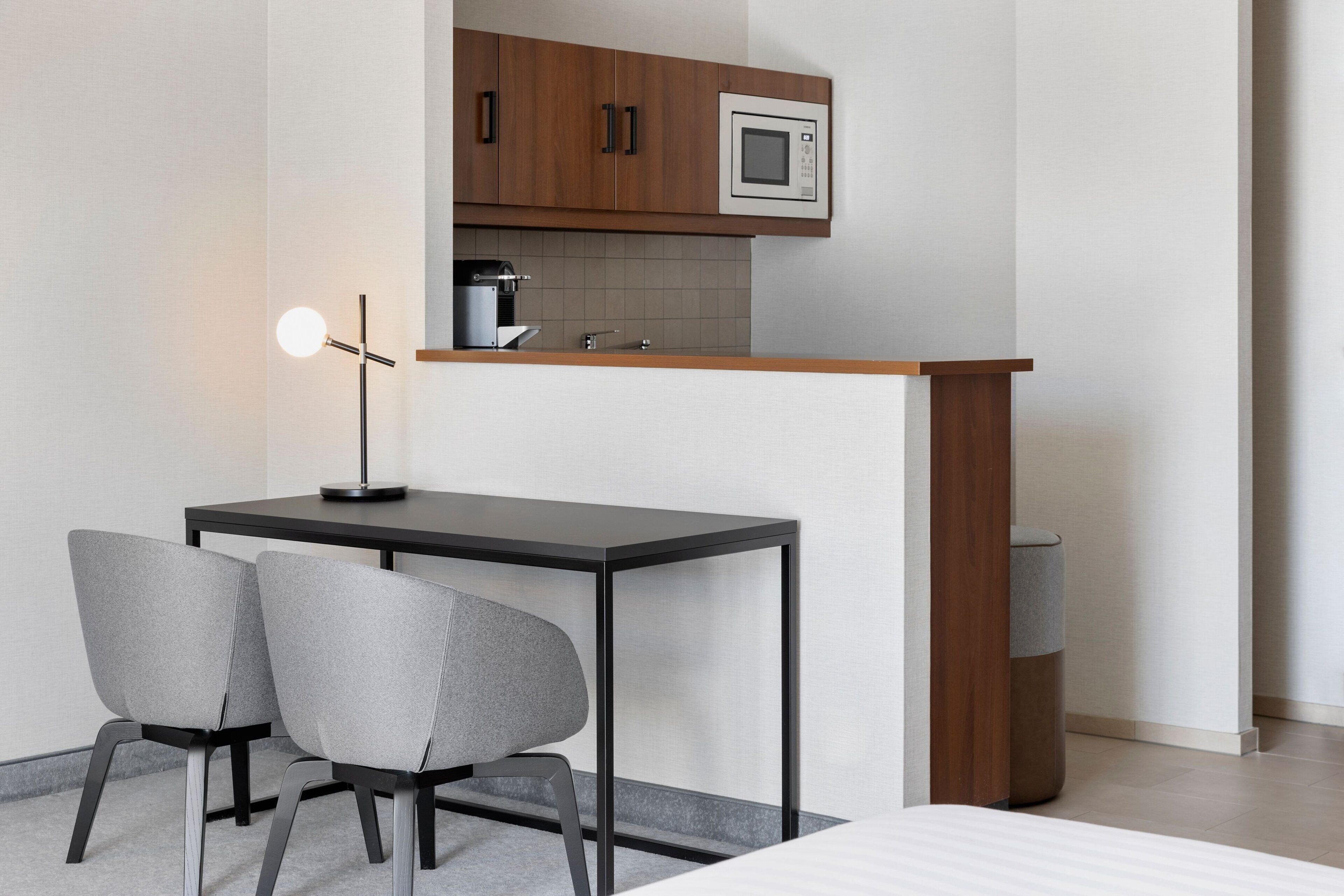 Een kleine keuken met een magnetron, een koffieapparaat en een houten aanrecht. Er staat een klein tafeltje met twee stoelen en een vloerlamp.