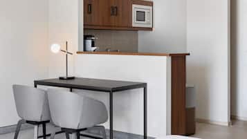 Een kleine keuken met een magnetron, een koffieapparaat en een houten aanrecht. Er staat een klein tafeltje met twee stoelen en een vloerlamp.