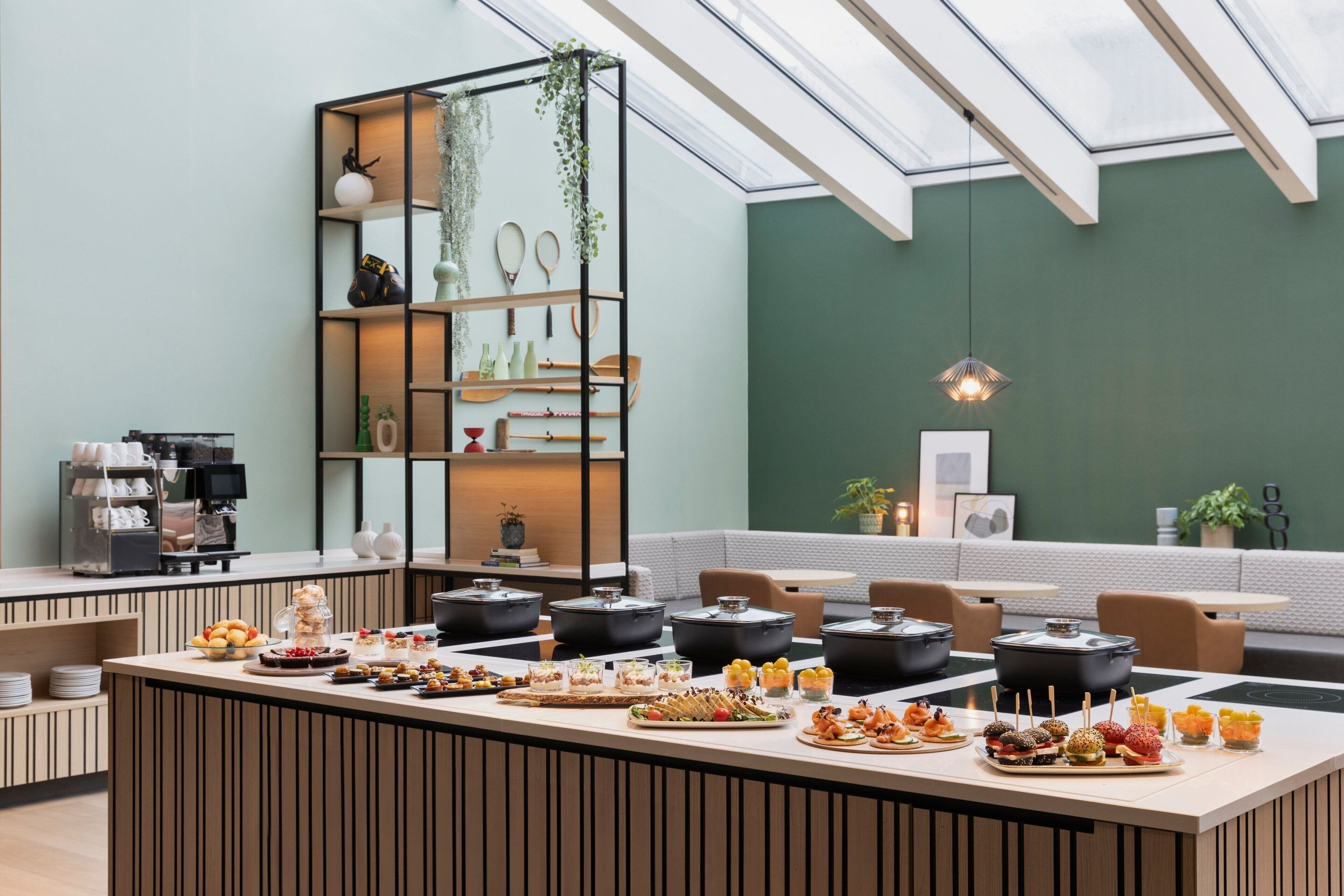A modern dining area with a central island featuring a variety of food items, surrounded by seating and a green wall with a skylight.