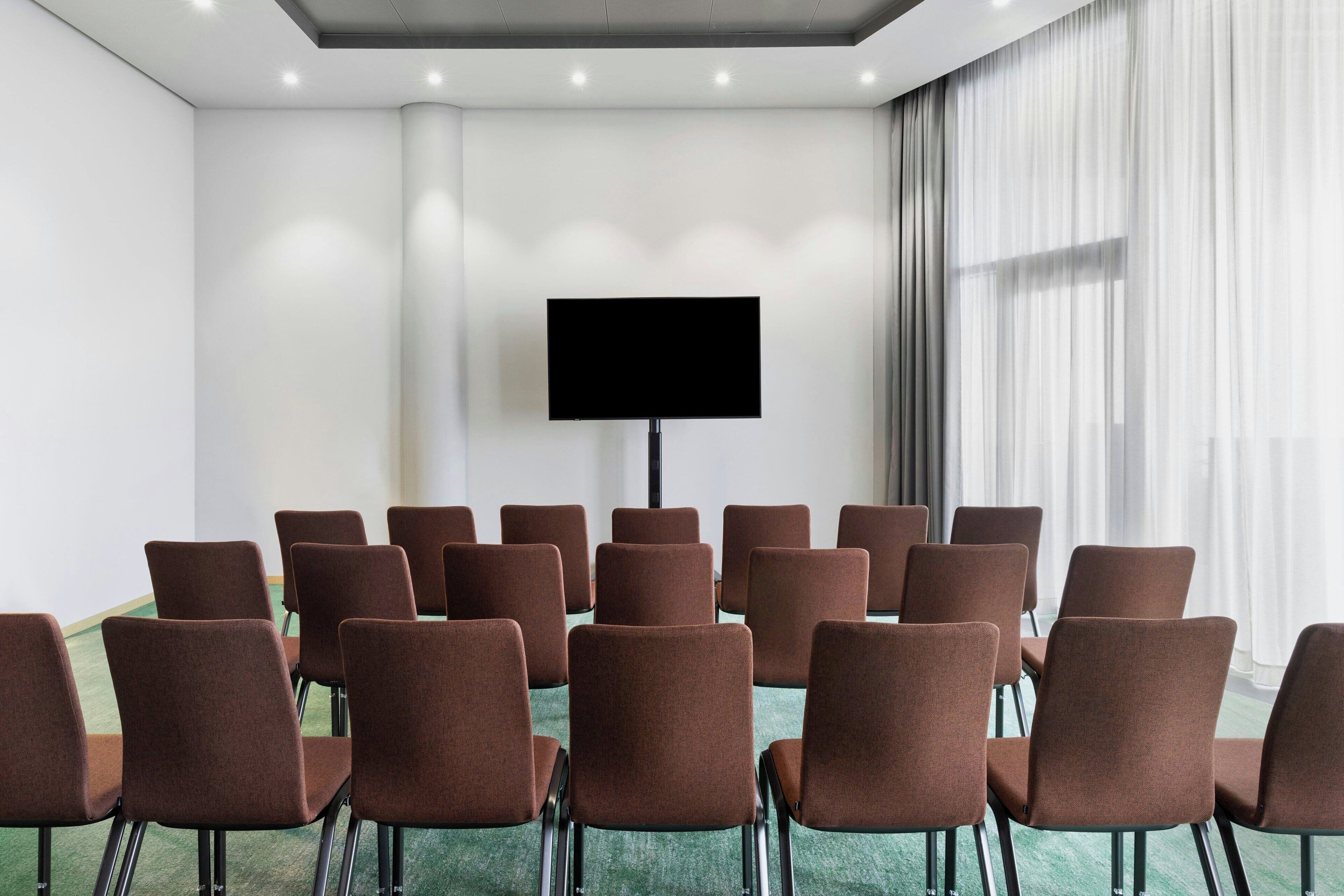 A conference room with rows of brown chairs, a large flat-screen TV on the wall, and a large window with sheer curtains.