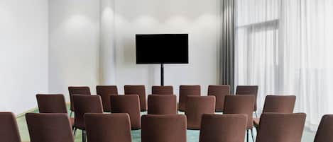 A conference room with rows of brown chairs, a large flat-screen TV on the wall, and a large window with sheer curtains.