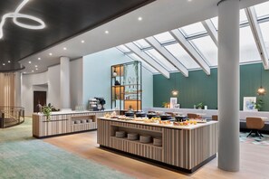 A modern kitchen with a central island, a coffee station, and a seating area with a view of the sky through skylights. - Courtyard by Marriott Basel (Pratteln)